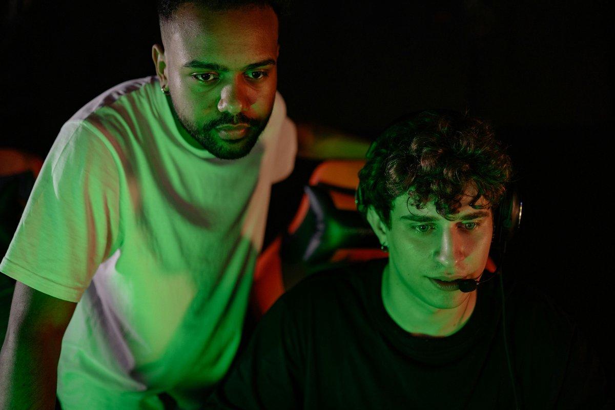 Two focused gamers lit by green screen light, one leaning in beside a teammate wearing a headset