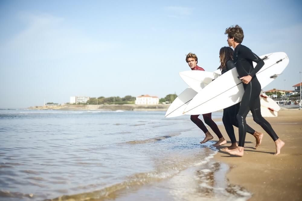 Three young men in wetsuits carrying surfboards along the shoreline.