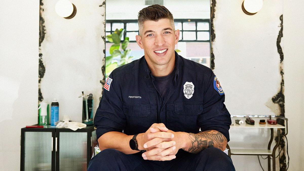A man wearing a firefighter's duty shirt, posing for a photo from a barberchair
