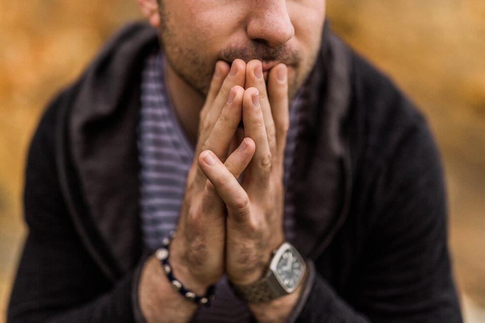 A man sits with his hands pressed together against his mouth, looking down, in a pose that suggests worry or deep thought.