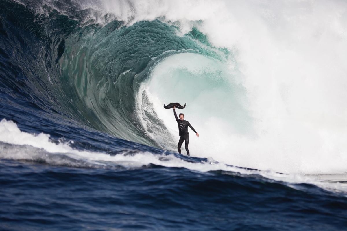 A man is surfing a wave and holding a giant moustache in the air