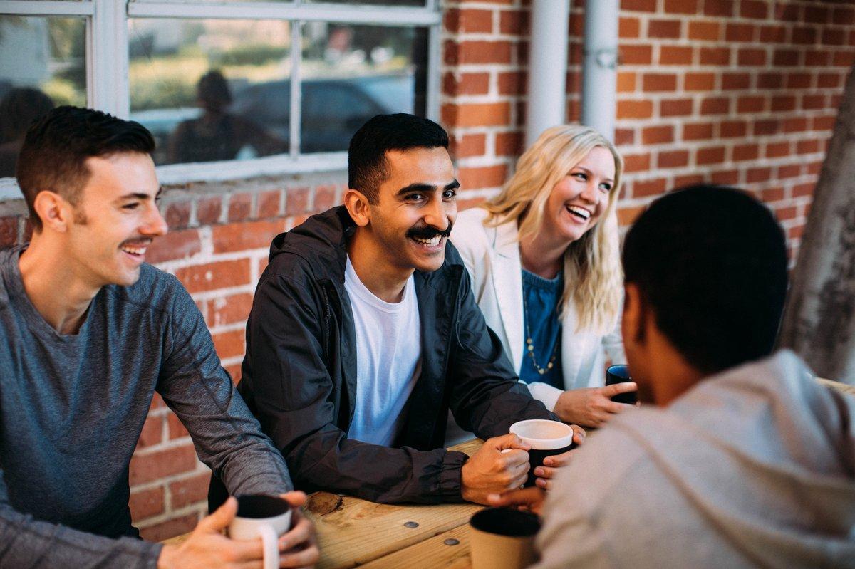 A group of four friends laughing and talking together over coffee at an outdoor table.