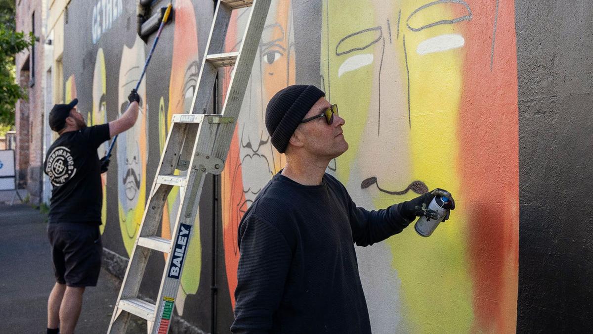 Street artist stands holds a spray paint can and looks at mural on wall, which he is painting.