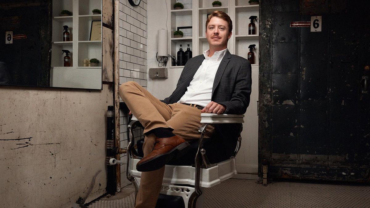 A suited young man sits in the barberchair of a New York City barbershop