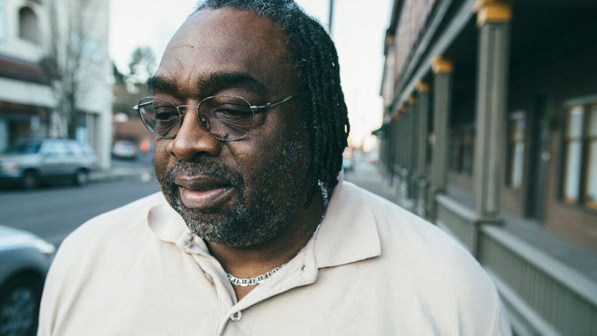 Black man with short dreads and glasses standing on the sidewalk