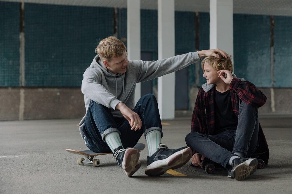 Two young men sitting on skateboards, one ruffling the other's hair while they talk.