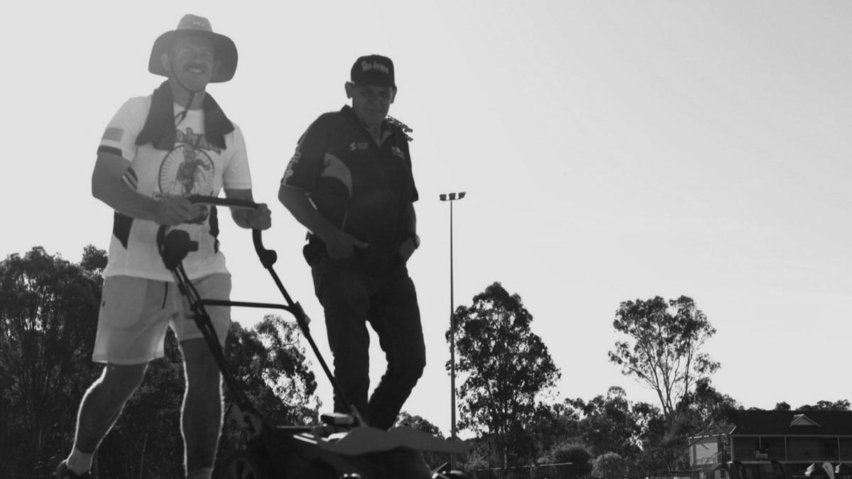 Two men walk in a park, one pushes a lawnmower, the other walks by his side. One looks at the camera with thumbs up. The other looks at the ground.