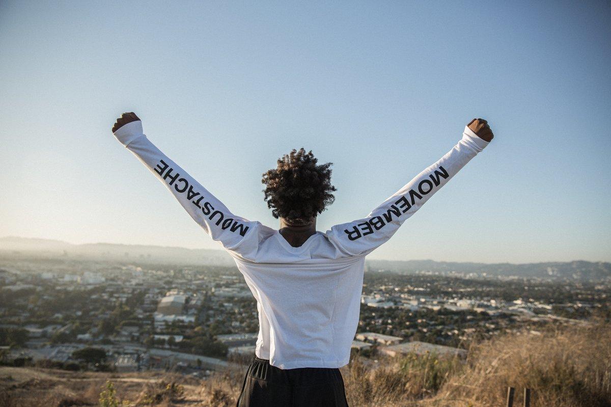 A man wearing a Movember long-sleeve shirt stands on a hilltop with arms raised, looking out over a city skyline.