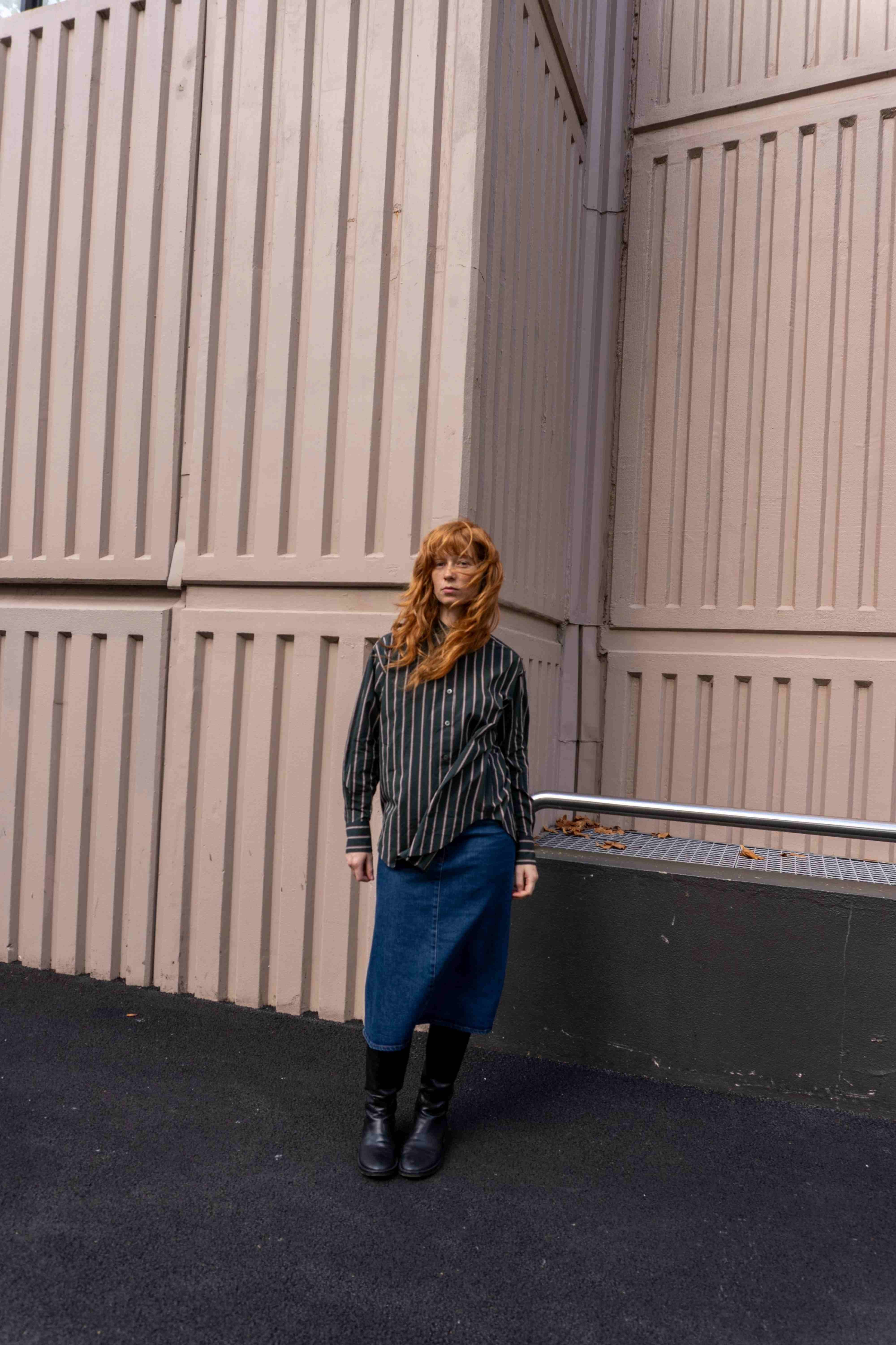 Model with red hair wearing a green shirt and denim skirt in front of a pink building