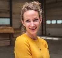 Vrouw met krullend haar en een gele trui, glimlachend in een lichte binnenshuisomgeving.