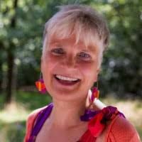 Vrouw met kort blond haar, glimlachend buiten met groene bomen op de achtergrond.