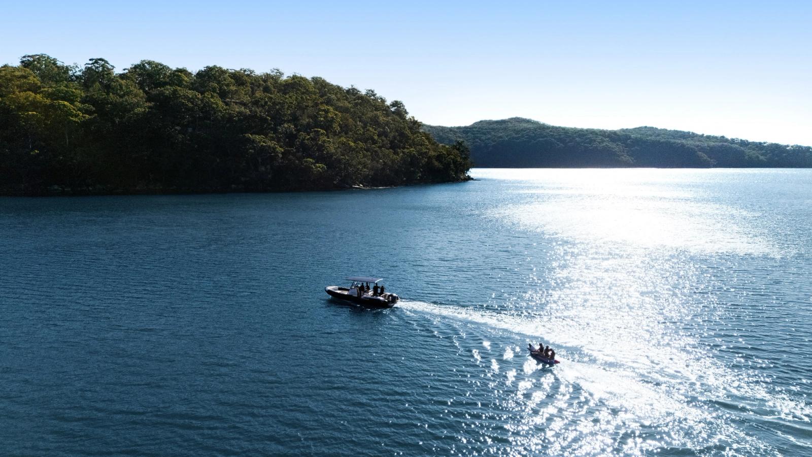 Aerial view of tenders crossing sunlit water past a wooded island headland