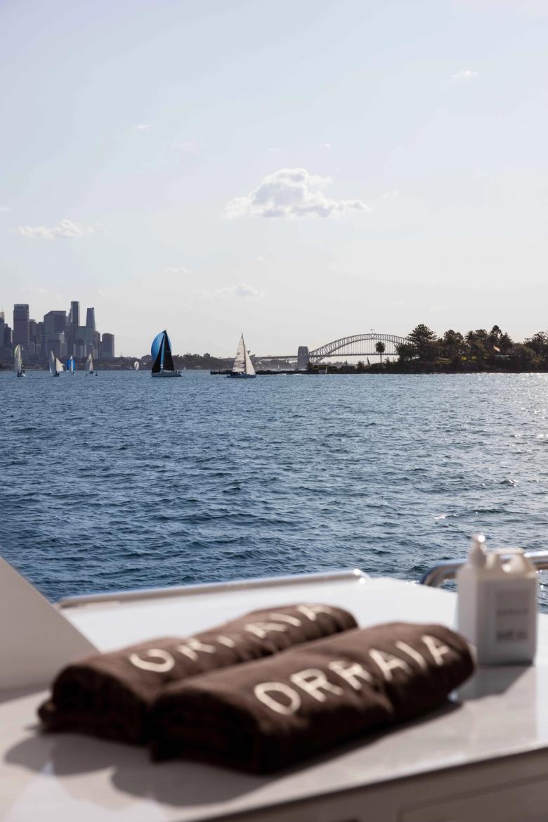 ORRAIA branded towels on deck with sailboats and the Harbour Bridge behind