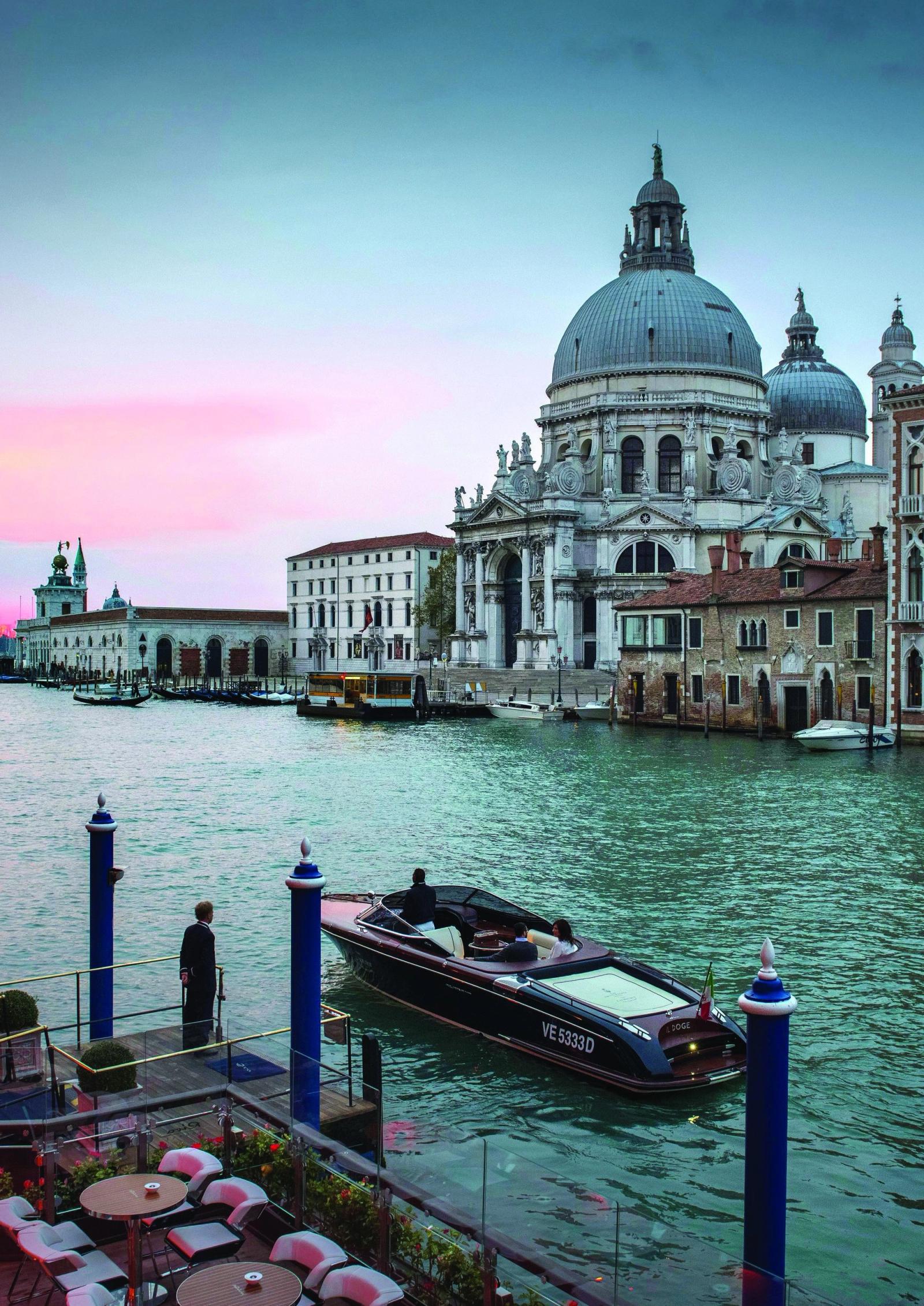 A Riva runabout on a Venetian canal at dusk beneath the domes of Santa Maria della Salute, the sky washed pink