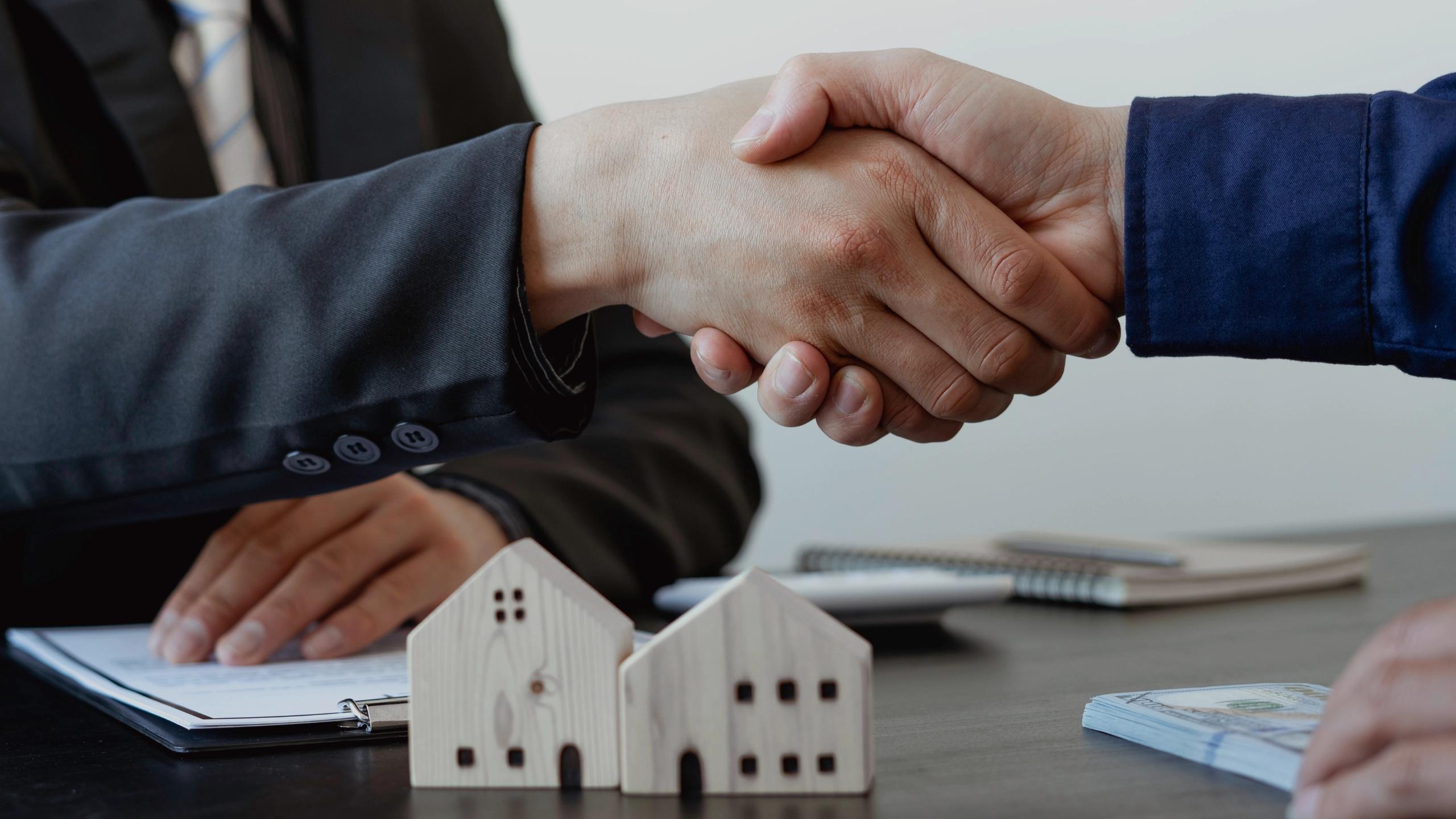 Two people shaking hands over property documents with small house models on the table.
