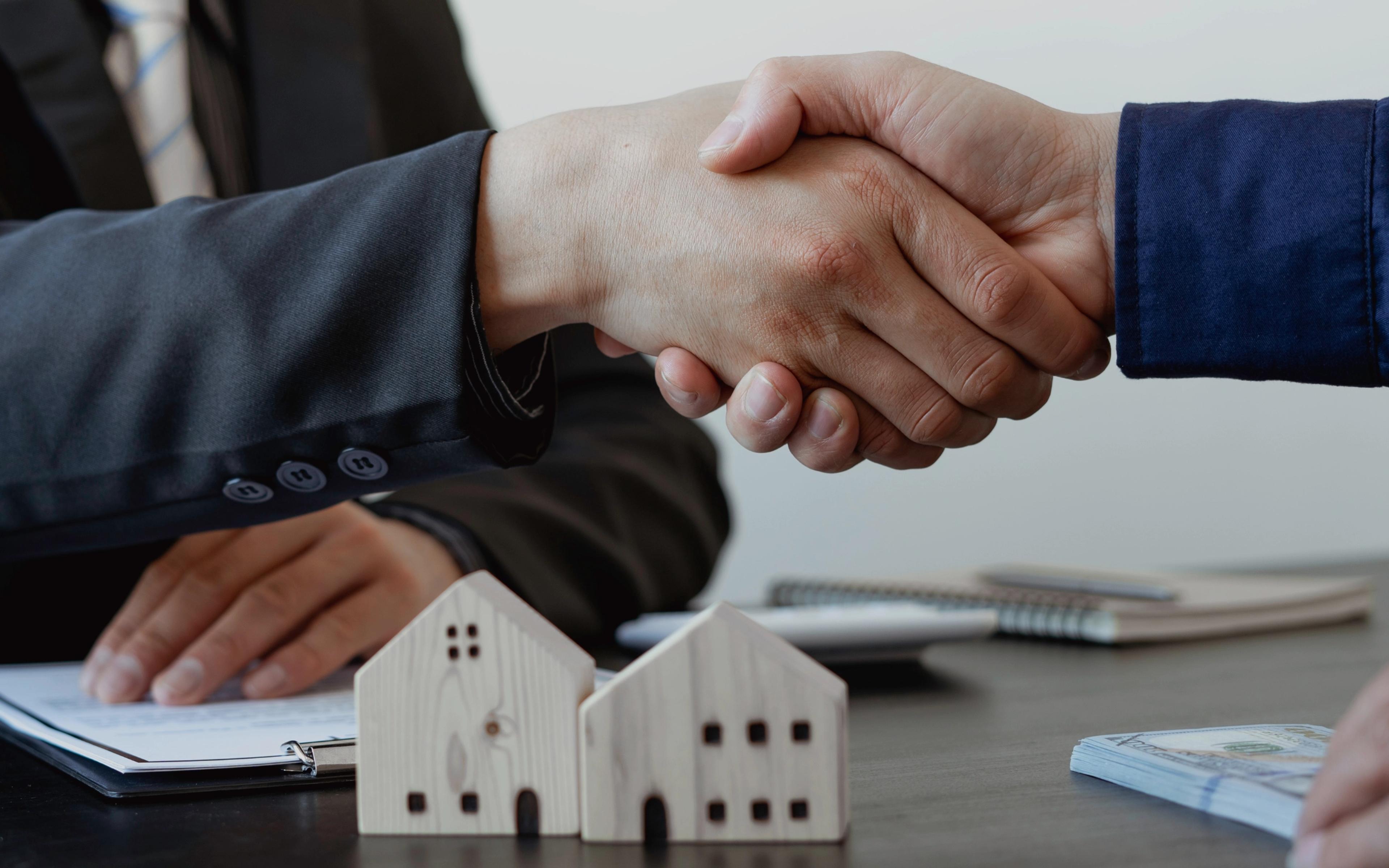Two people shaking hands over property documents with small house models on the table.
