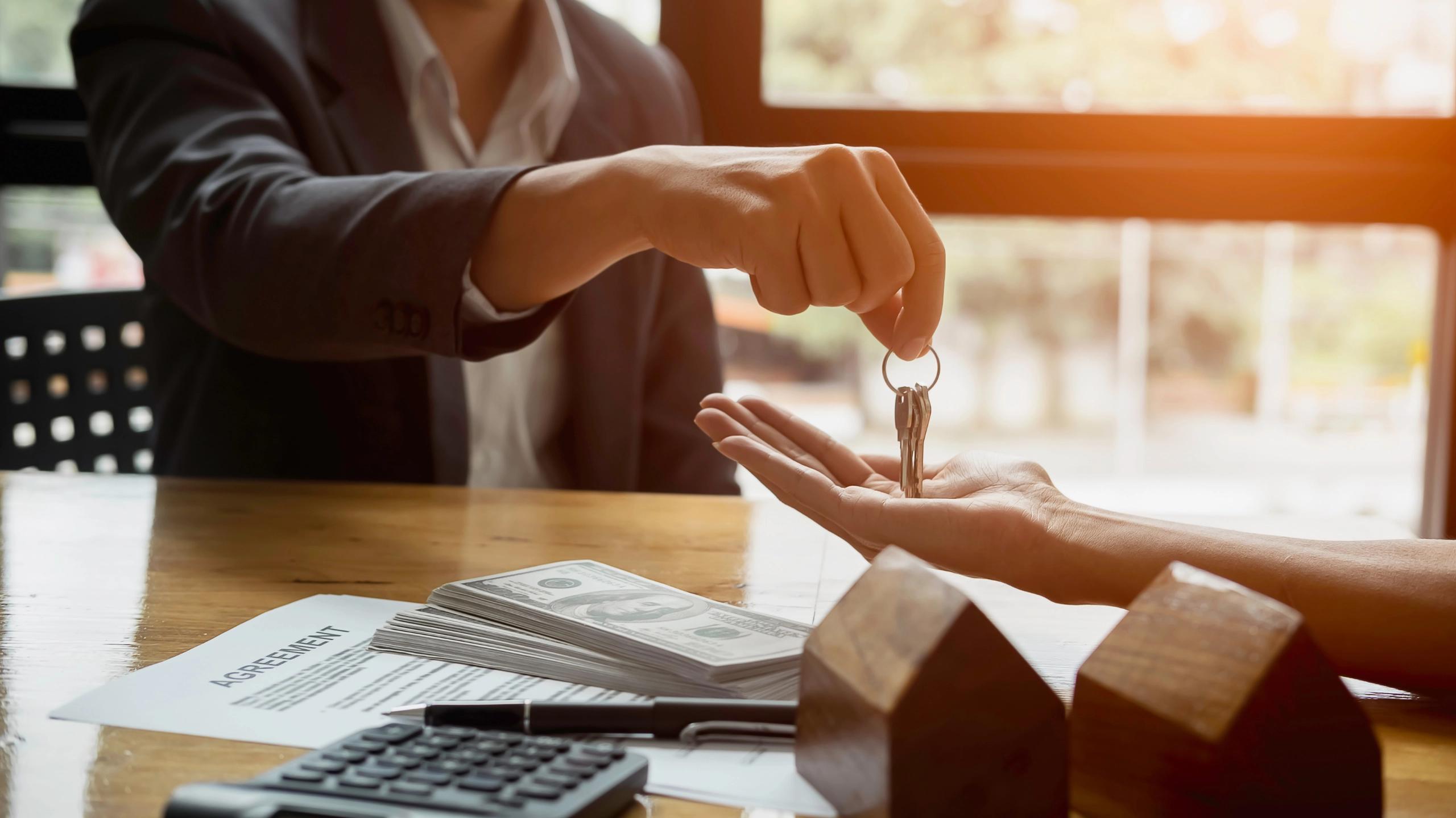 A person hands over a set of keys to another person at a table with cash and a contract.