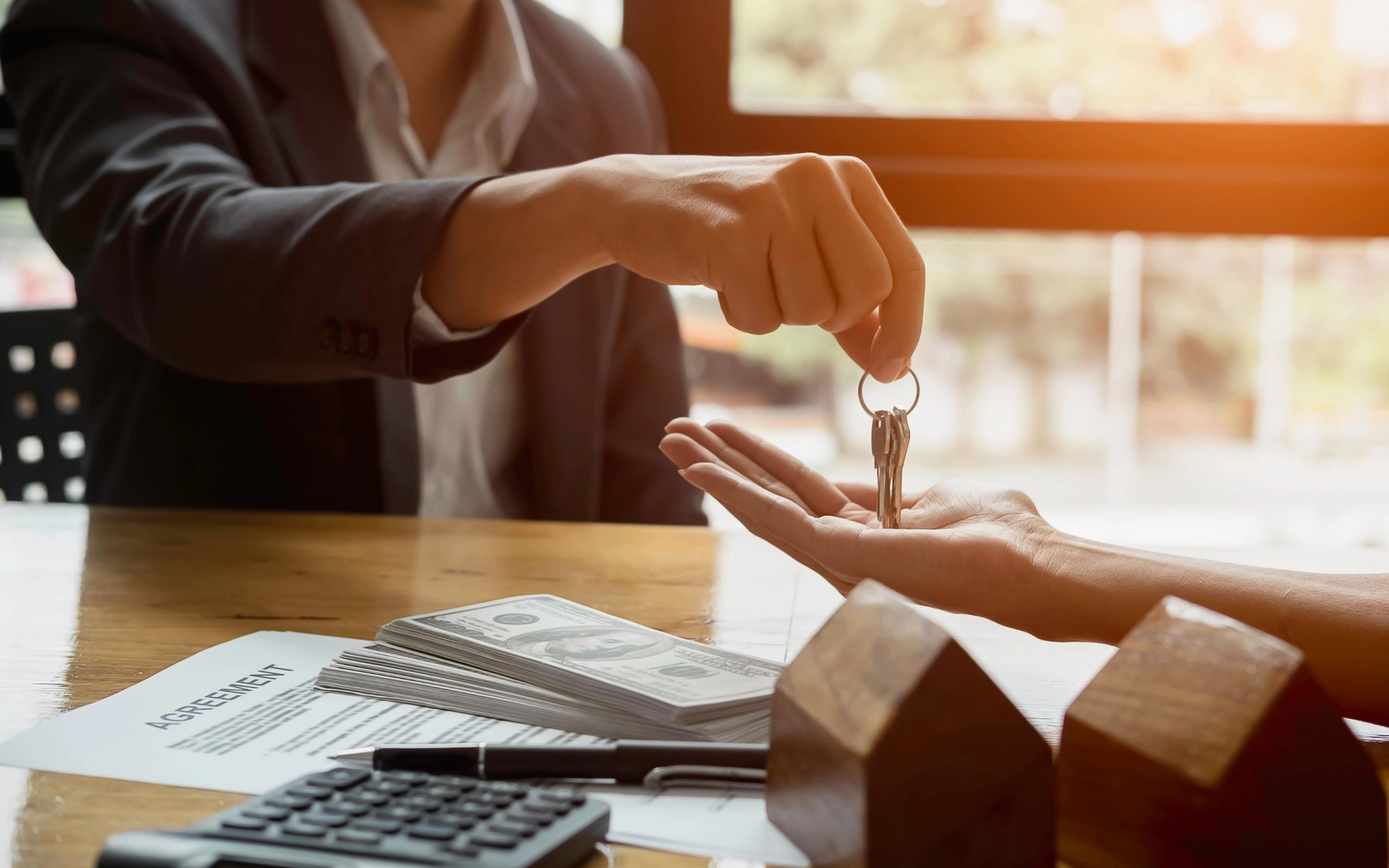 A person hands over a set of keys to another person at a table with cash and a contract.