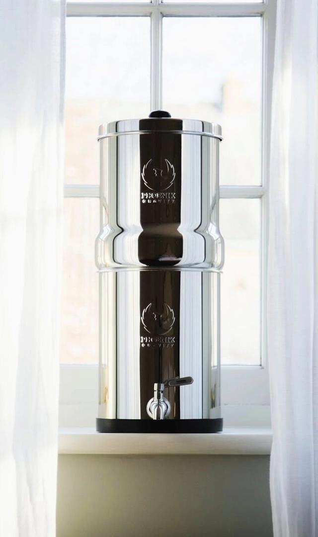 Stainless steel water filter sits on a windowsill, featuring a spout and a logo for "Phoenix Clarity."