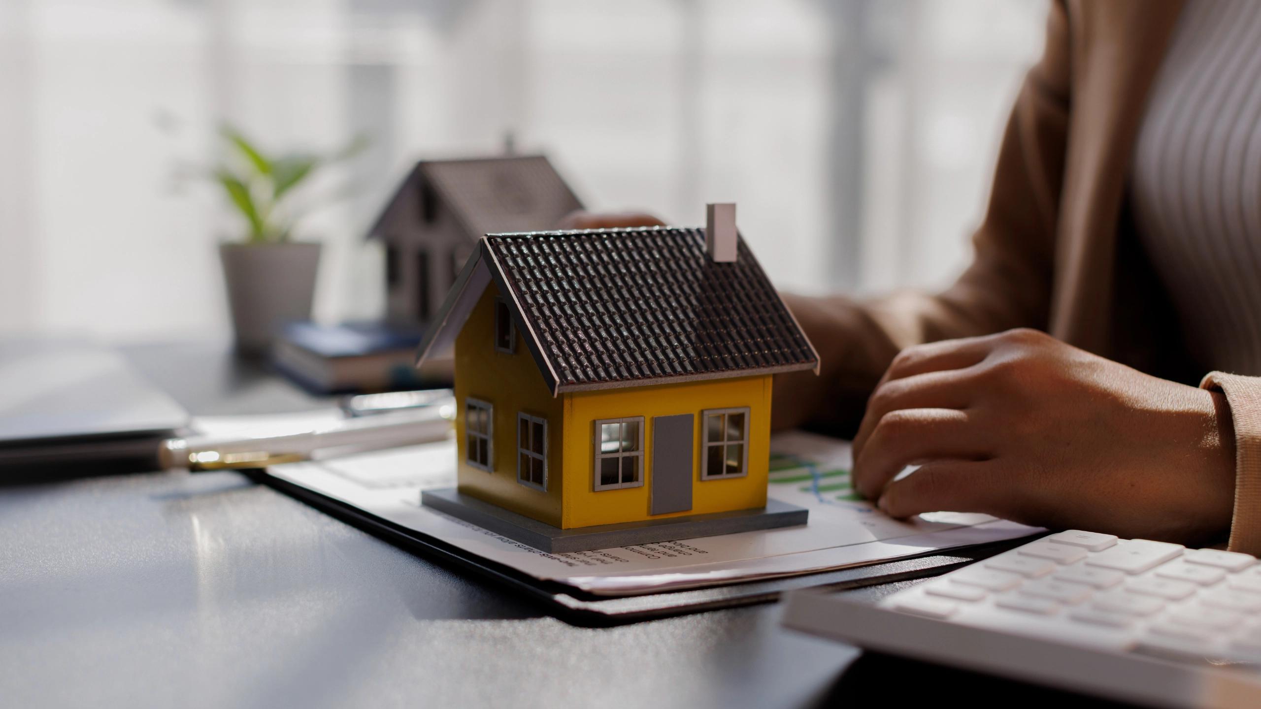 Small house model on a desk beside documents and a person working.