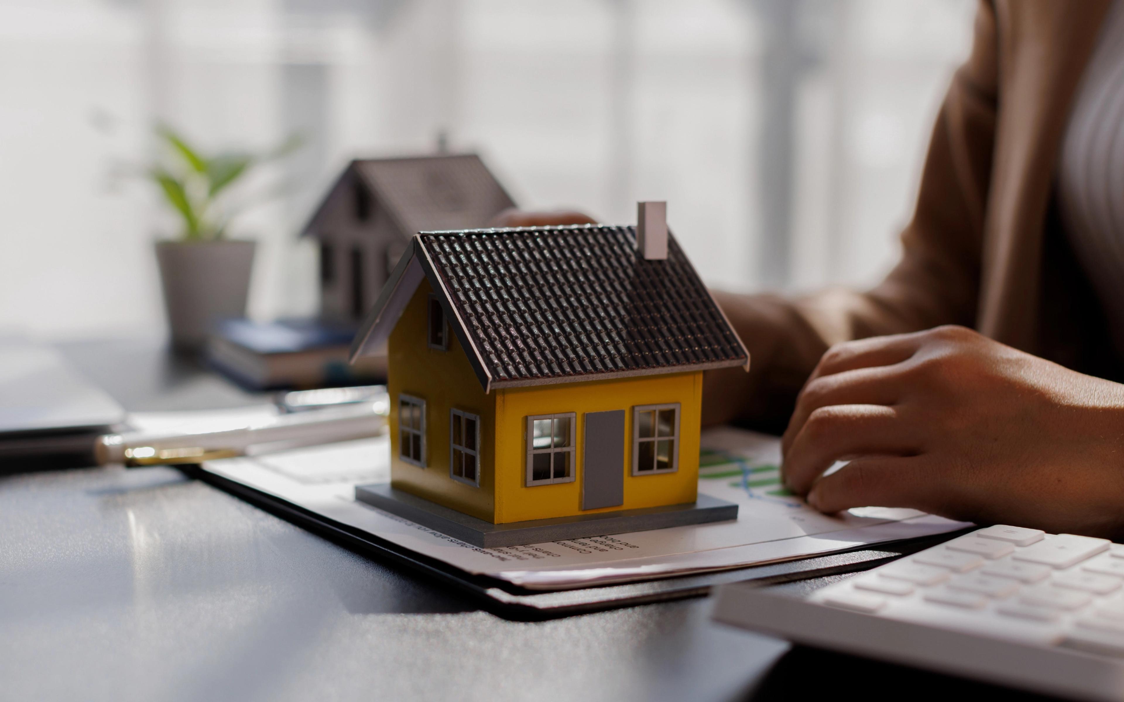 Small house model on a desk beside documents and a person working.