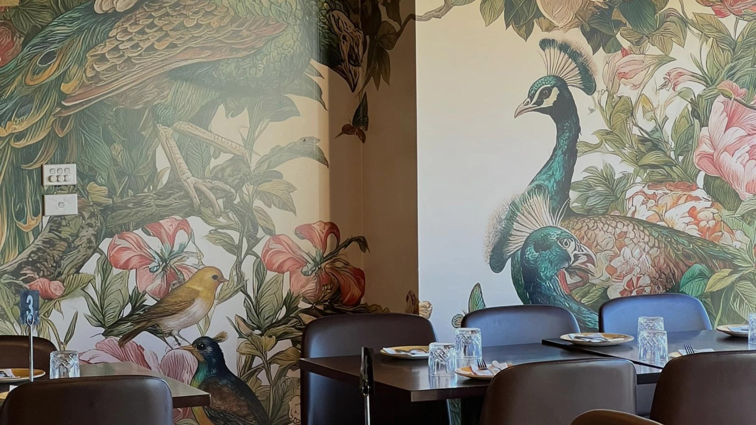 Restaurant interior with colourful peacock and bird mural on the wall and tables set for dining.