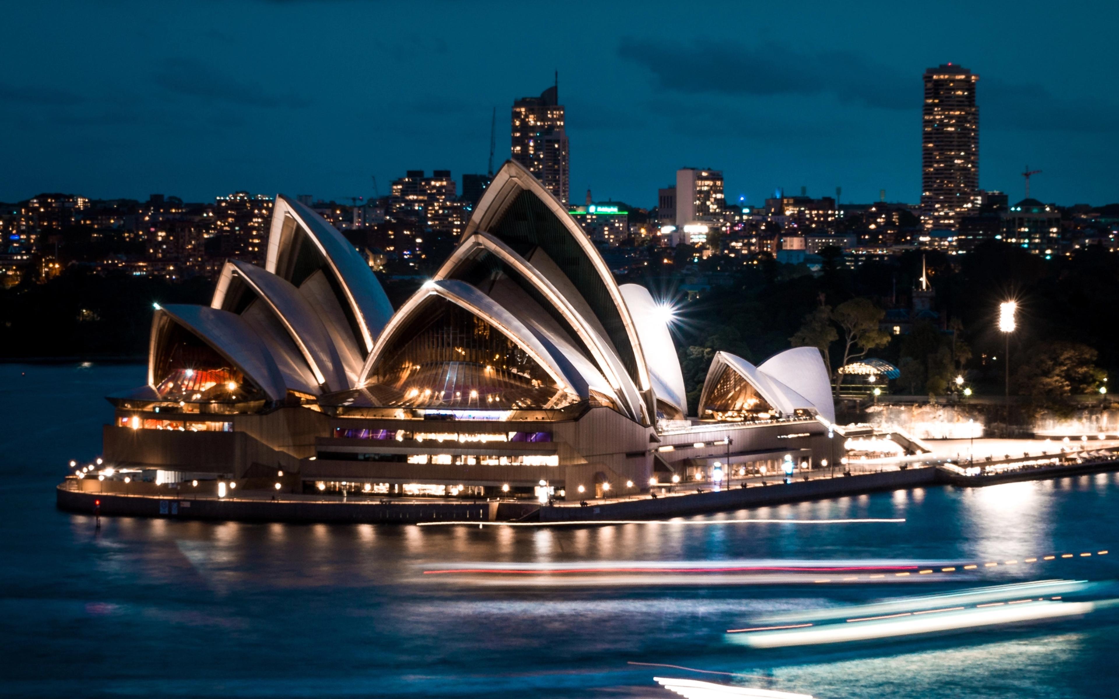 Sydney Opera House illuminated at night beside harbor with light trails and city skyline