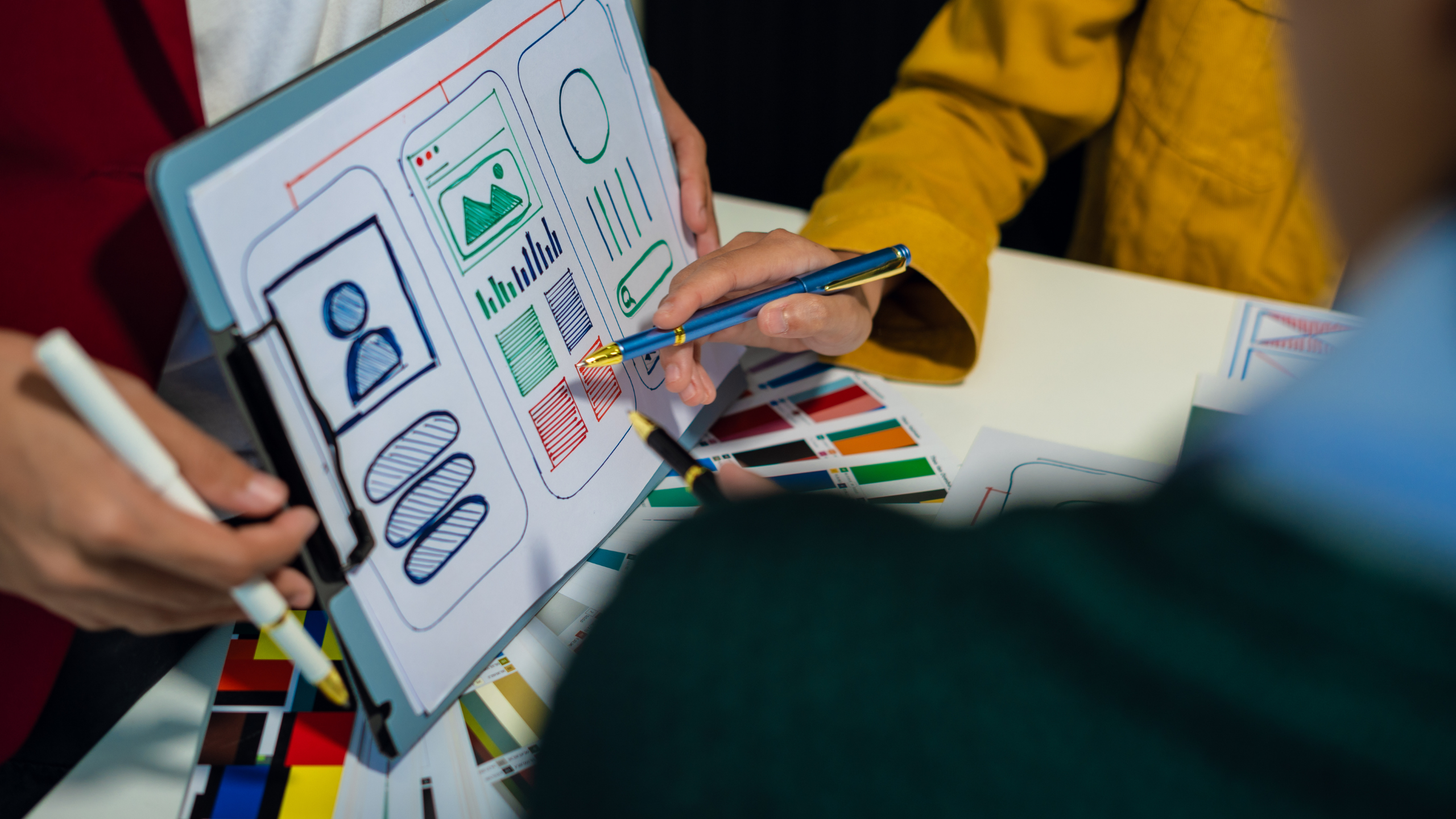 Designers reviewing a website or mobile app wireframe on a tablet, discussing layout elements and color selections with printed color swatches on the table.