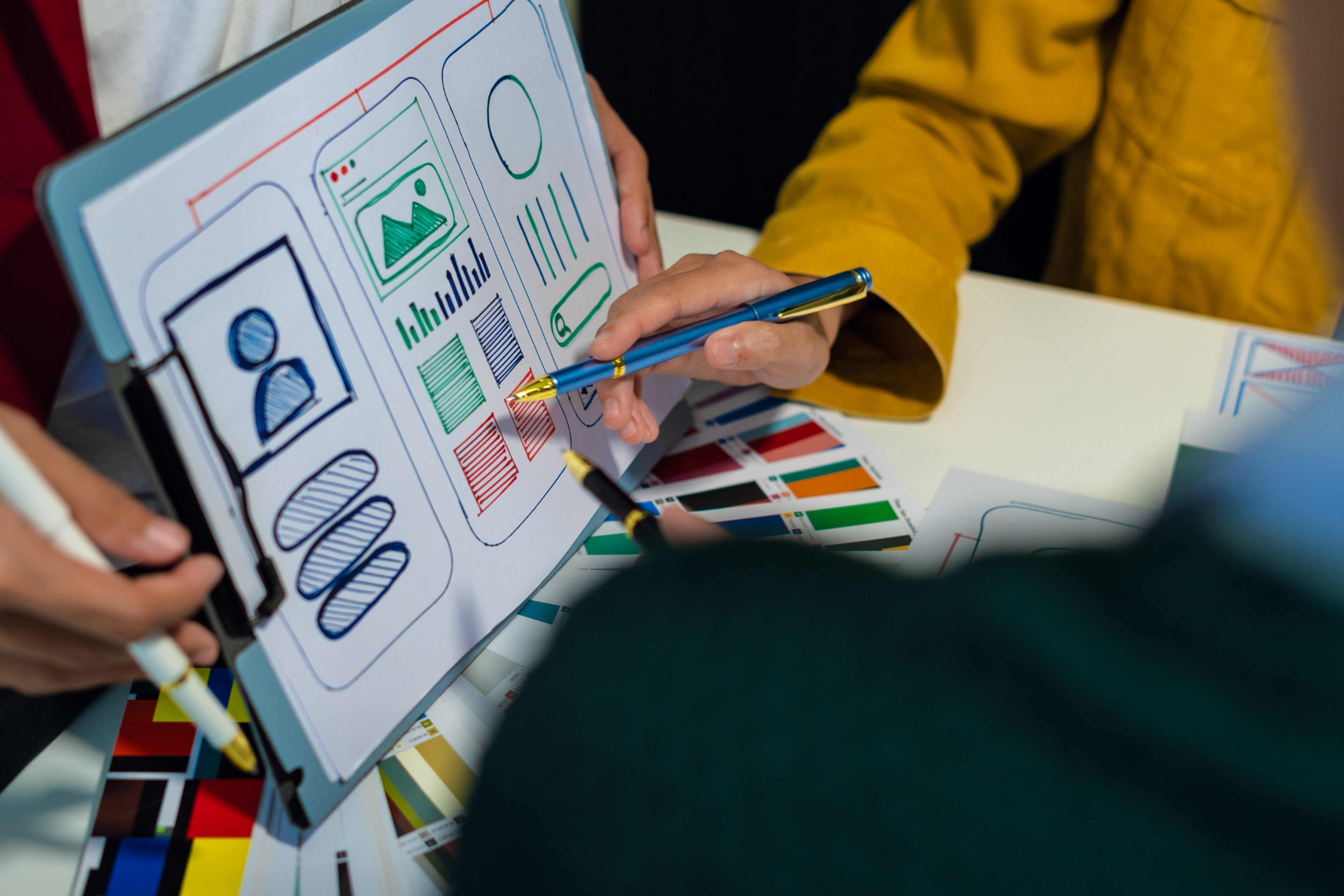 Designers reviewing a website or mobile app wireframe on a tablet, discussing layout elements and color selections with printed color swatches on the table.
