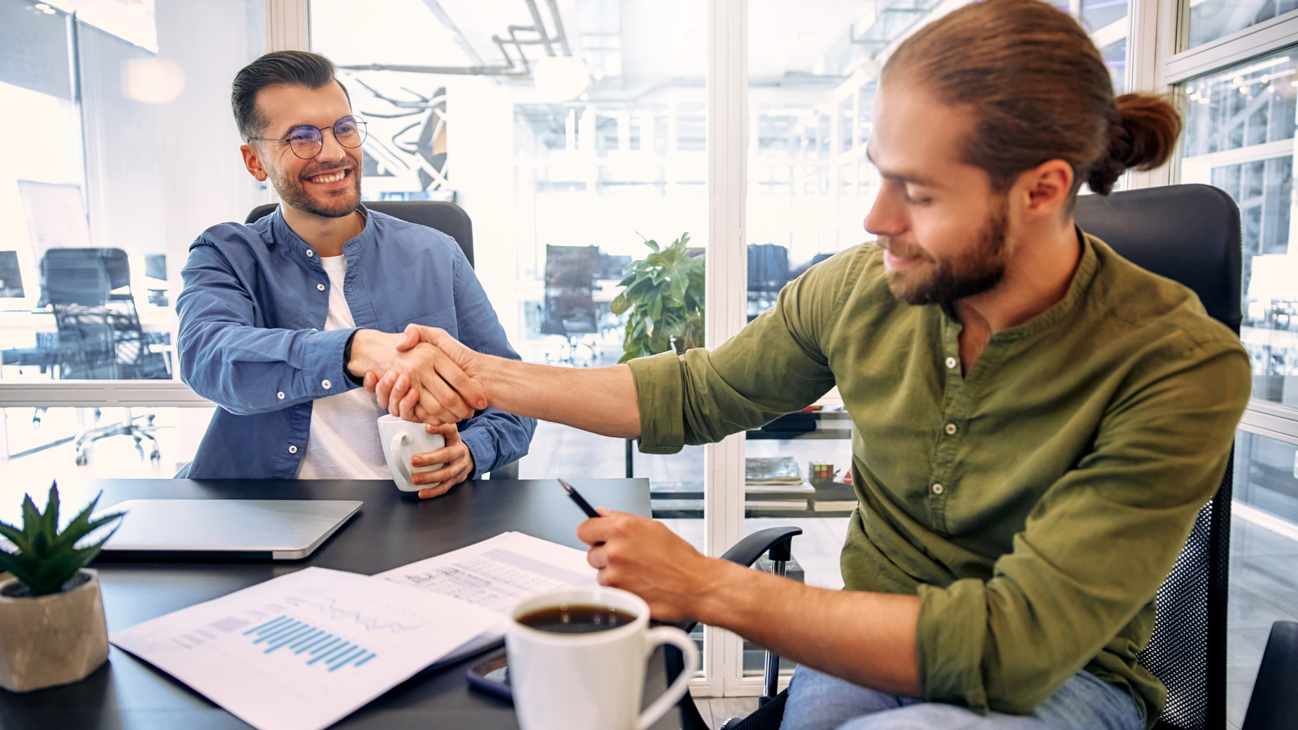 Two professionals shaking hands across a desk in a modern office, representing trust, partnership and business agreement.
