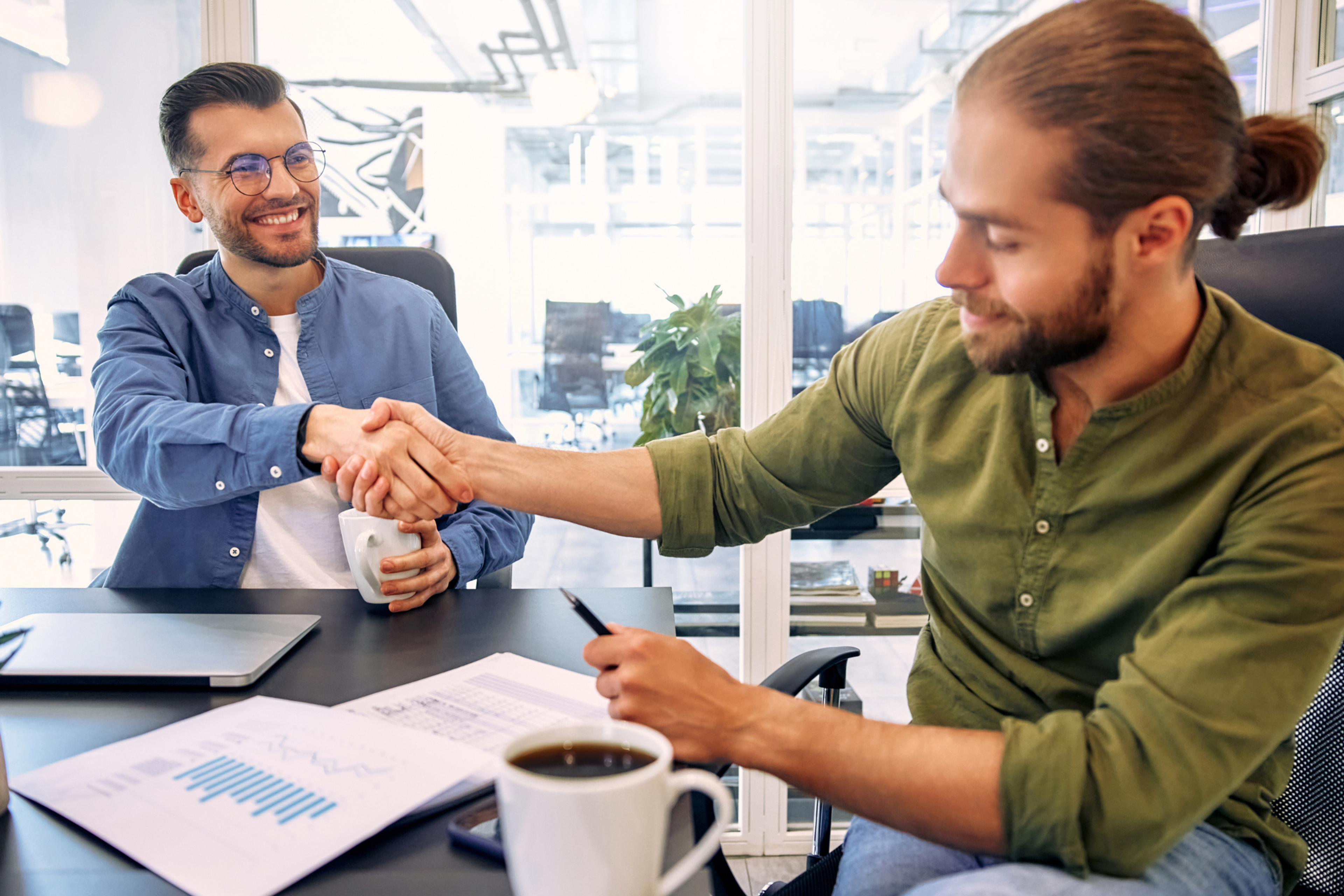 Two professionals shaking hands across a desk in a modern office, representing trust, partnership and business agreement.