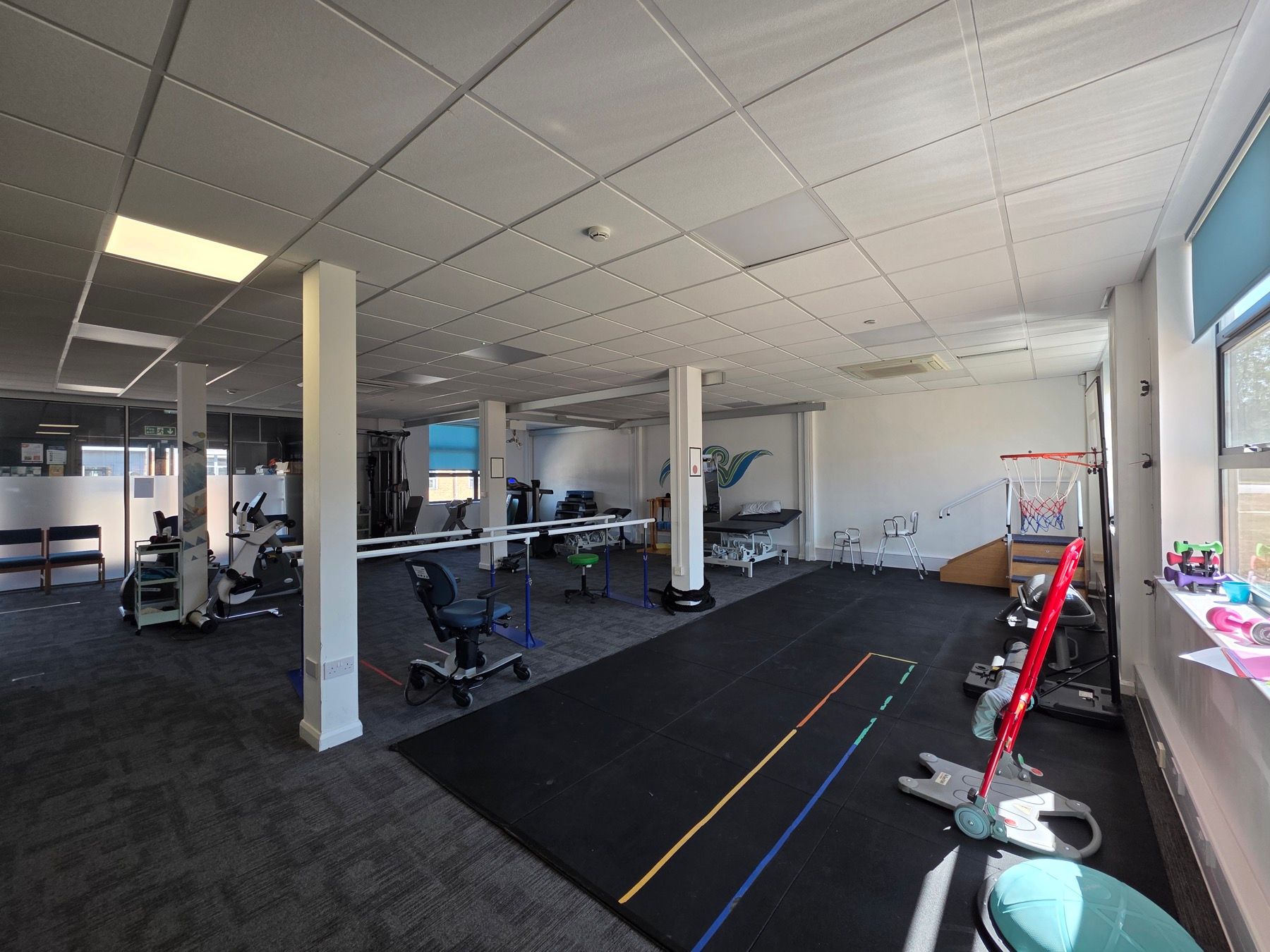 A wide view of the rehabilitation gym: parallel bars, treadmill, exercise bikes and an overhead hoist track in an open, step-free space