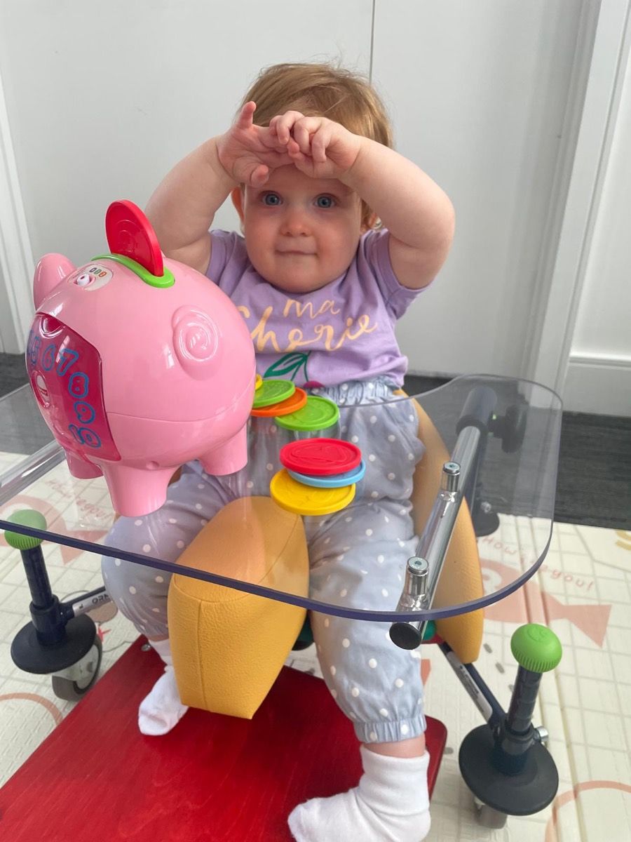 A baby sitting supported in a standing frame with a play tray, reaching towards stacking toys