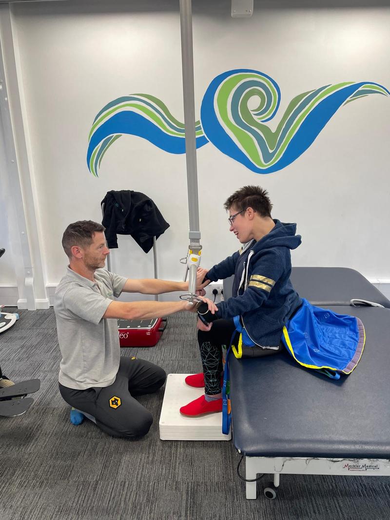 A physiotherapist kneeling to support a client practising standing at the edge of a treatment couch, with an overhead hoist strap for support