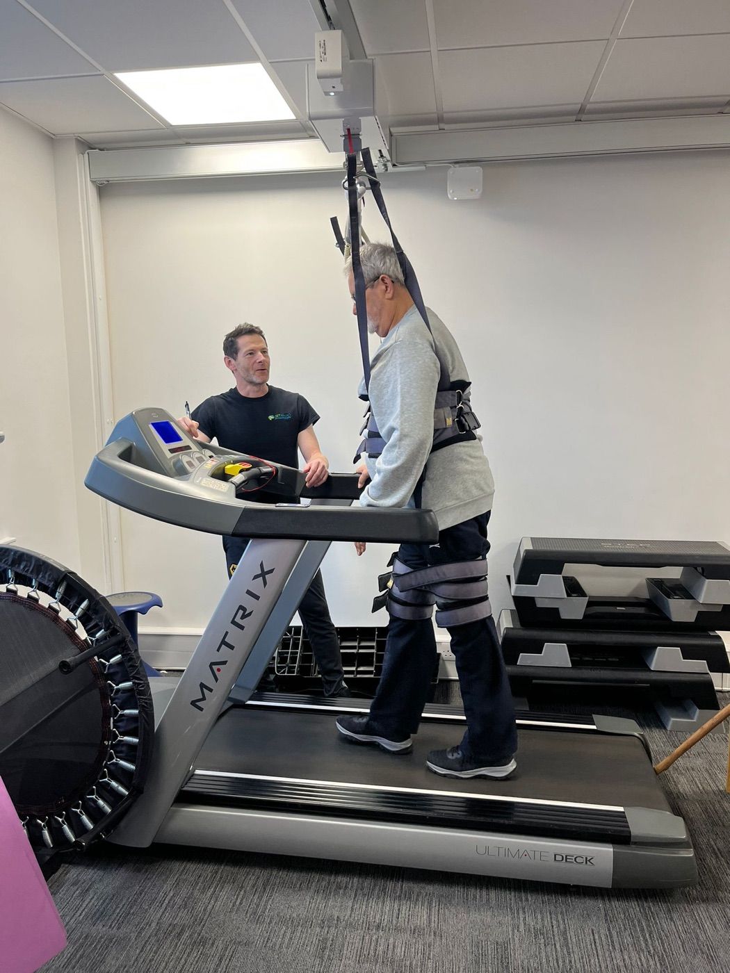 A client walking on the treadmill in a support harness while a physiotherapist assists