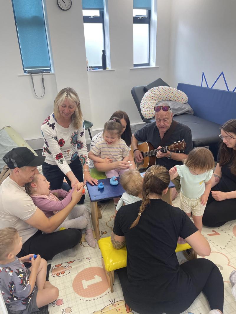 Children playing percussion at a low table while the music leader accompanies them on guitar