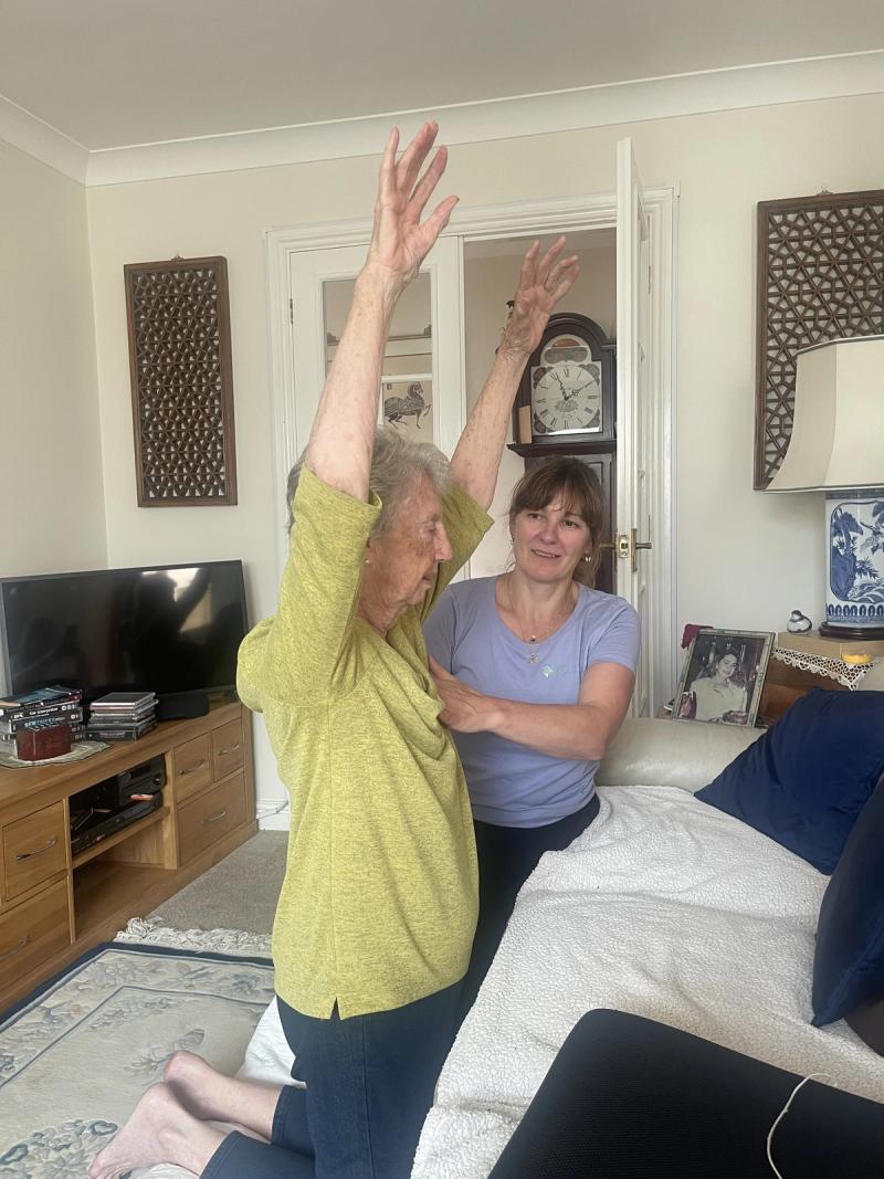 A client reaching both arms overhead with a physiotherapist supporting her, during a session at home