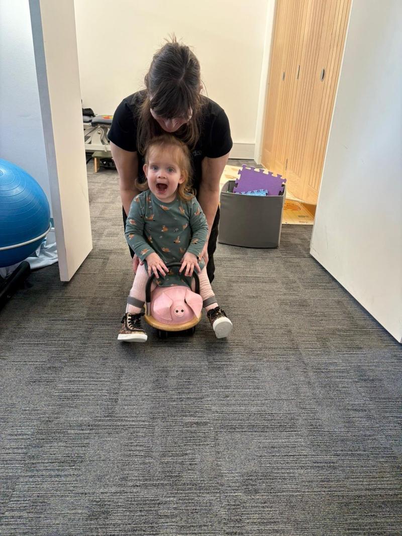 A physiotherapist steadying a smiling toddler on a ride-on toy in the clinic play area