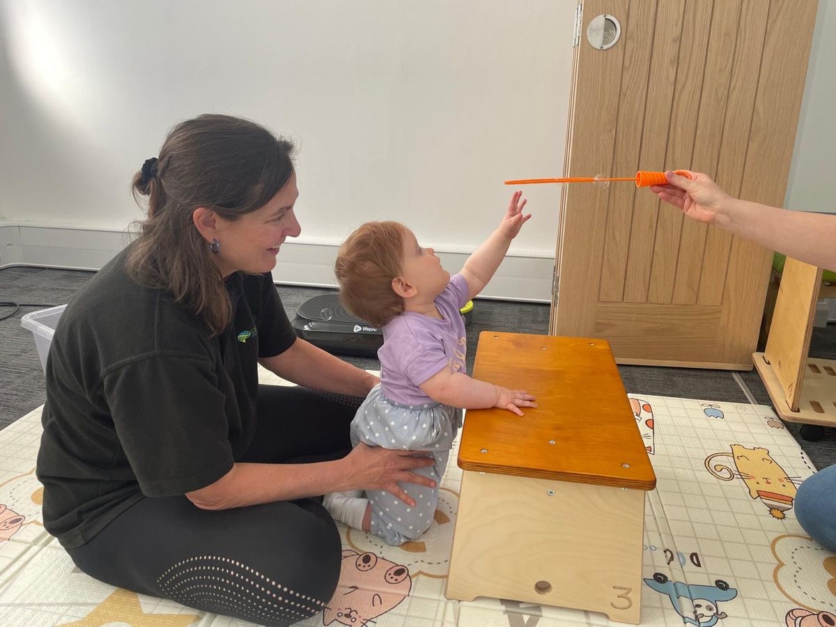 A physiotherapist on a play mat with a baby reaching towards a bubble wand