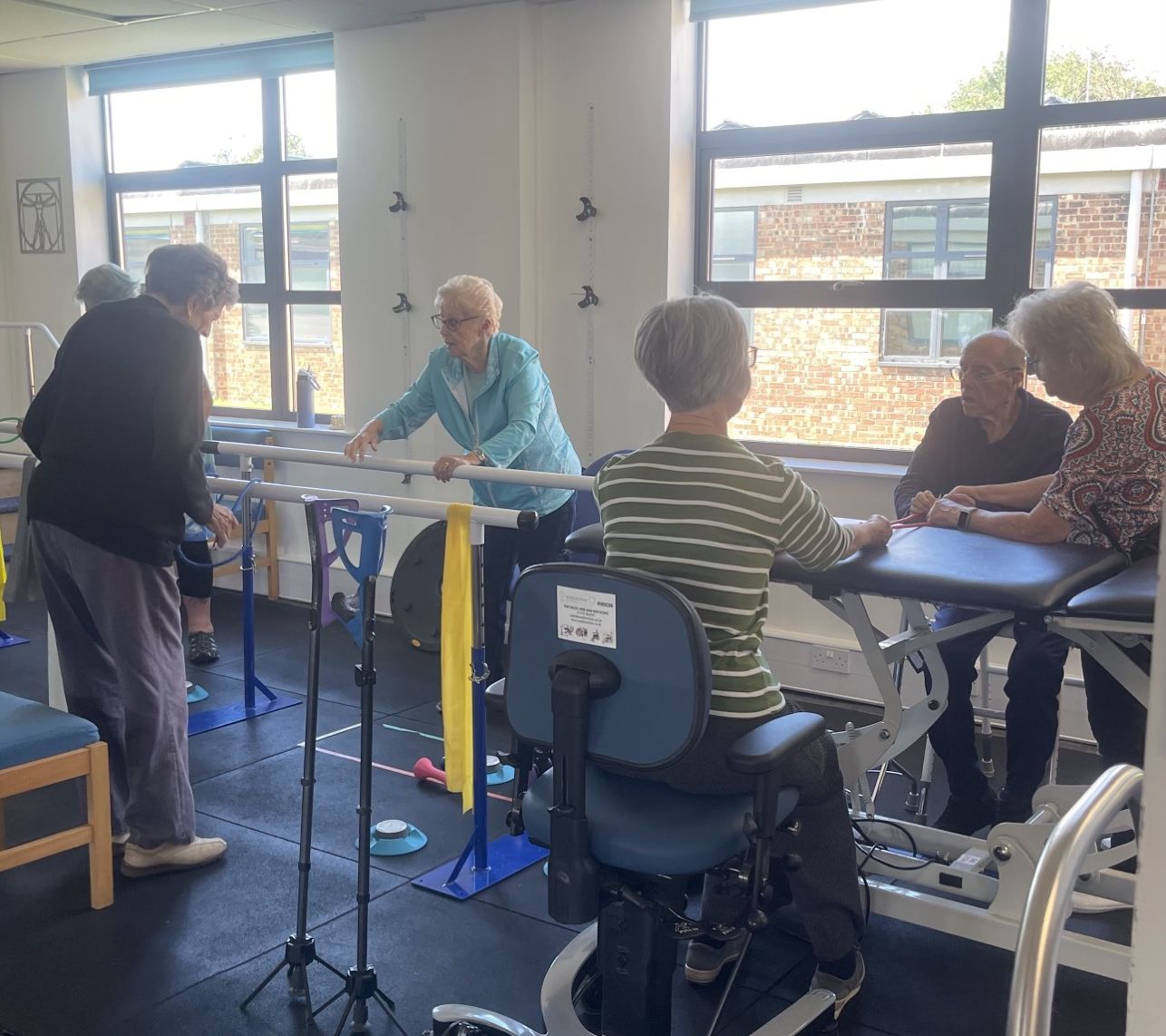 Group session in the clinic gym: one client walking at the parallel bars with a physiotherapist alongside, while others exercise seated at a plinth