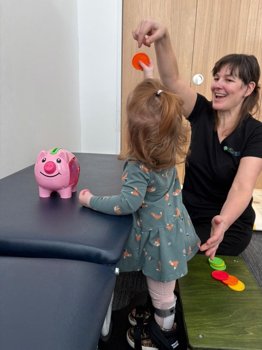 A toddler wearing an ankle splint, standing at a treatment plinth and posting shapes into a toy while a physiotherapist encourages her