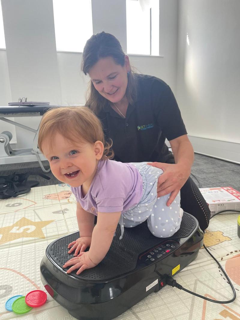 A baby taking weight through her arms on a vibration plate while a physiotherapist steadies her
