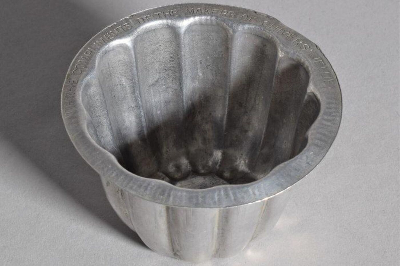 Miniature jelly mould, aluminium, flared shape with a flat rim and concave (on the inside) vertical grooves. Impressed letters around the rim `With the compliments of the makers of Chivers' jellies),