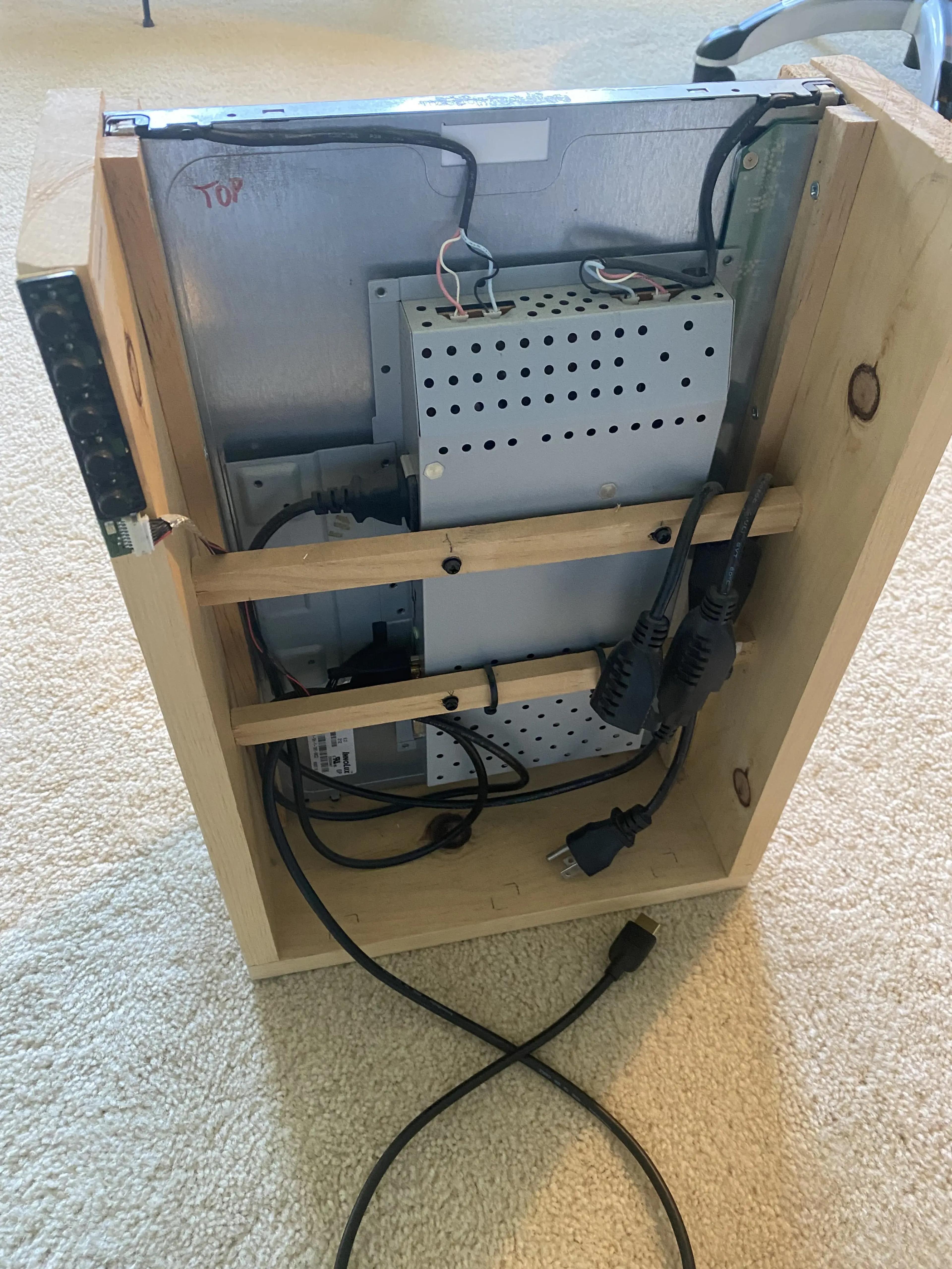 A shot of the back of the Magic Mirror. The wooden frame encloses the "brains" of the monitor. There are power cables and a HDMI cable coming out of the monitor. On the left side of the wooden casing is the monitor's buttons circuit board torn off and attached to the wooden casing.