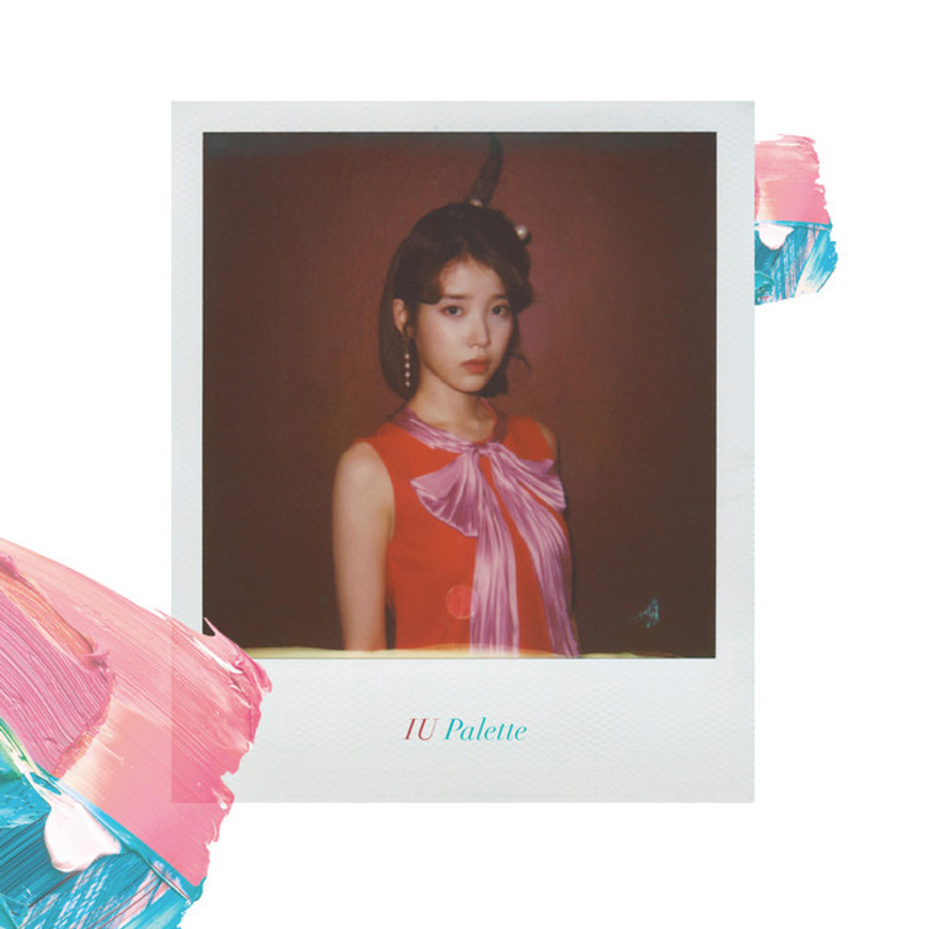 IU's 'Palette' album artwork, featuring a Polaroid-style photo of IU in a red dress with a pink bow, styled with her hair up. The Polaroid-style photo is labeled in a serif font: 'IU Palette' with 'IU' in pink and 'Palette' in cyan. Surrounding the photo are abstract strokes of pink and blue paint on the border of the frame.