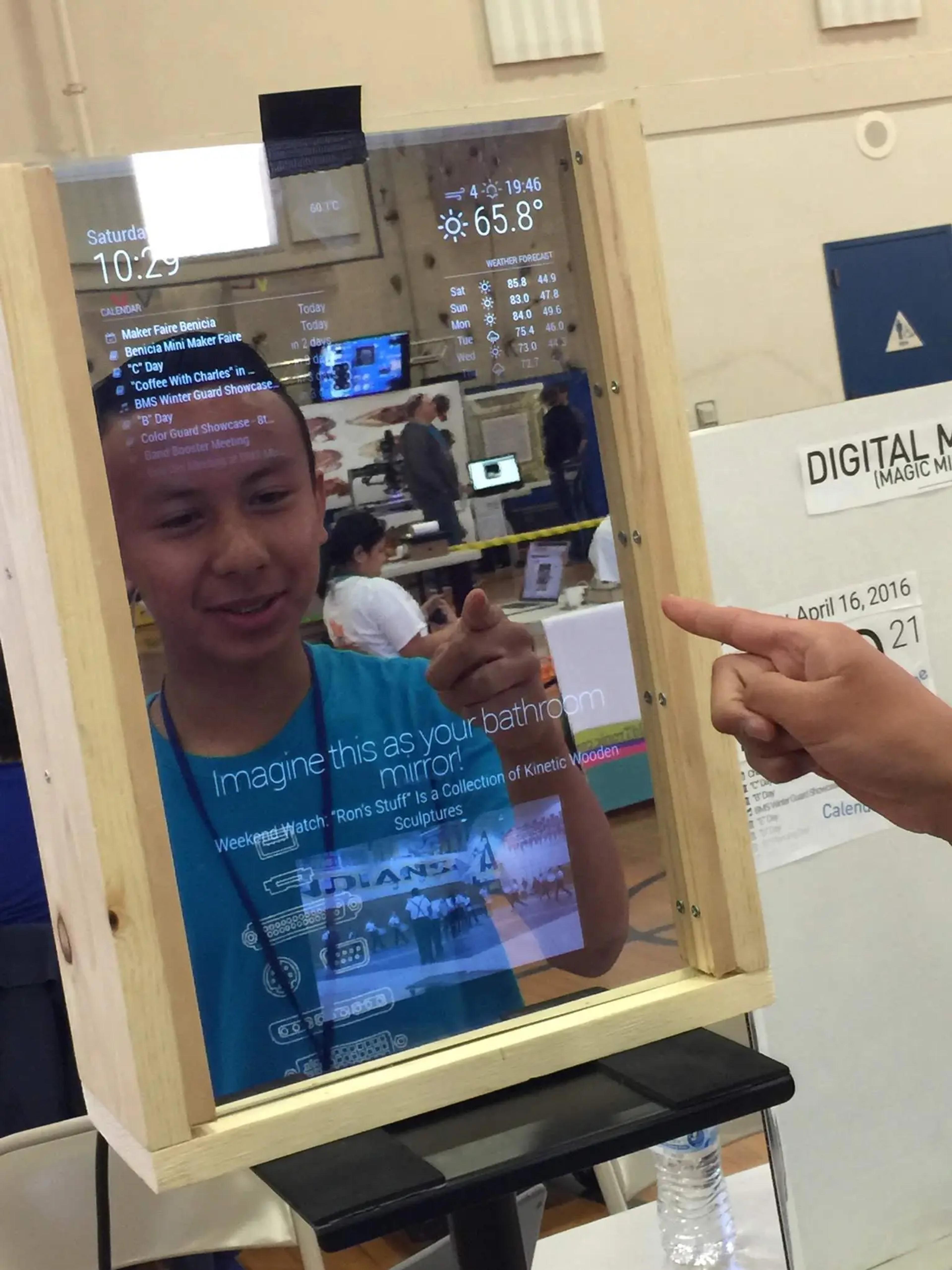 The Magic Mirror is elevated on a stand, about the height where someone can stand straight in front of it. In the reflection, you can see Kyle pointing at the mirror, along with the overlay of the various information showing "through" the mirror. Additionally, a video is playing towards the bottom of the mirror. Behind Kyle in the mirror's reflection, there are various stands and people. The stands are covered with various projects. Beside the mirror, the image partly shows a poster that says "Digital Mirror (Magic Mirror)", along with a screenshot of the Magic Mirror user interface, labeling what each section of the mirror shows.