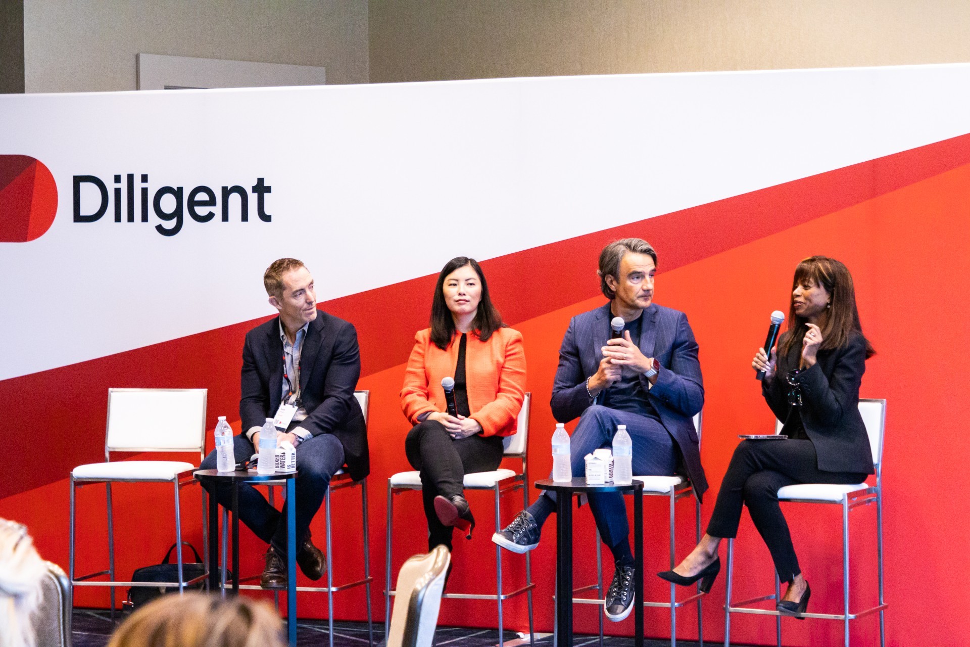 Panelists sit on stools upon a stage holding microphones and addressing an audience