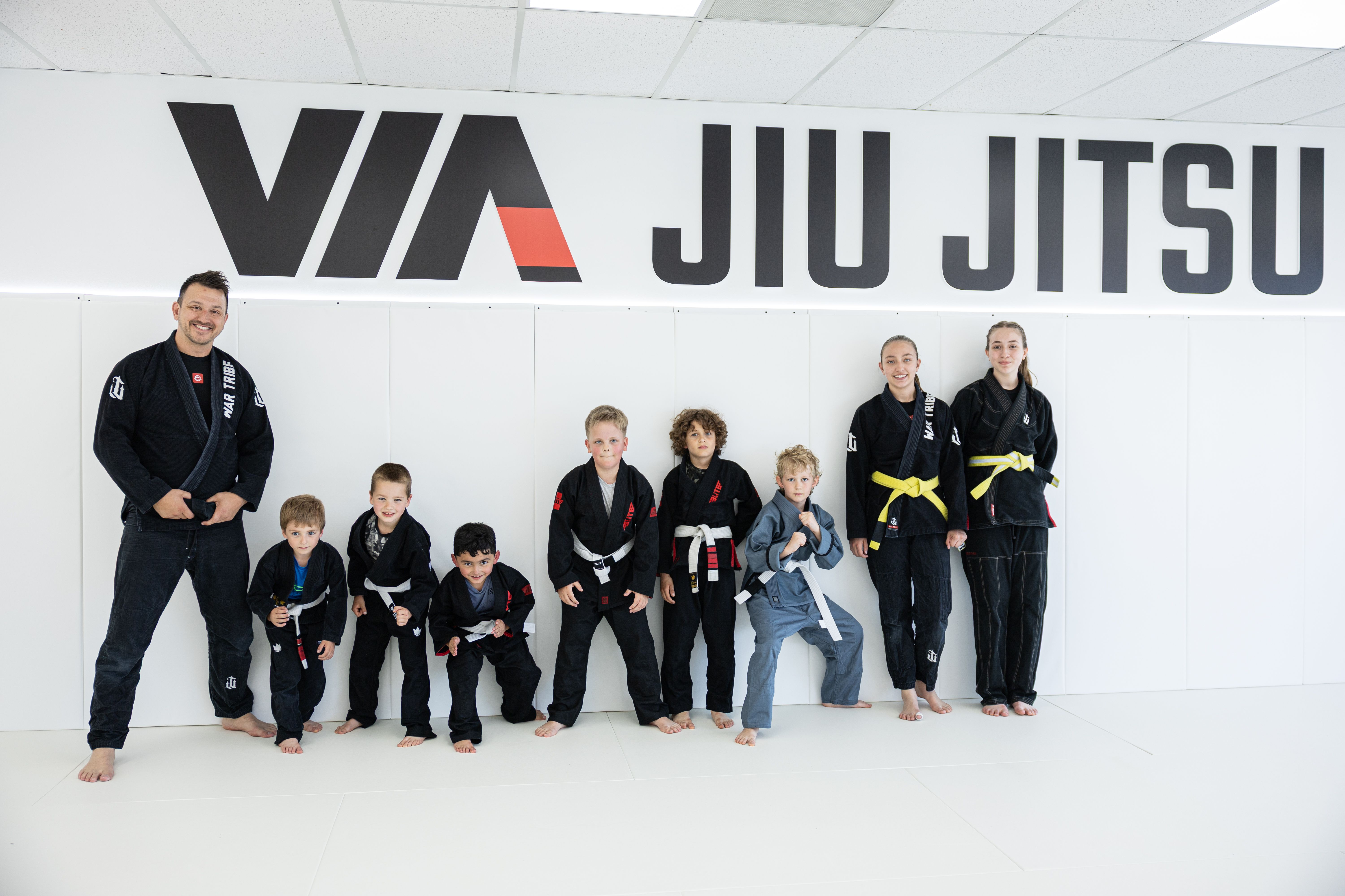 Kids and coach posing together under the Via Jiu Jitsu wall logo