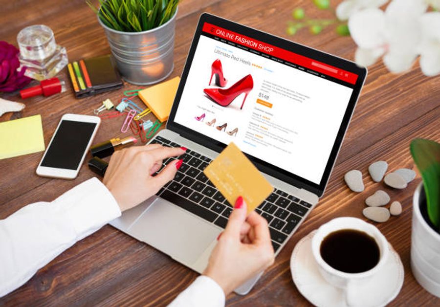 A woman with red nails holding a credit card while browsing a product page for red heels on an online fashion shop website displayed on a laptop