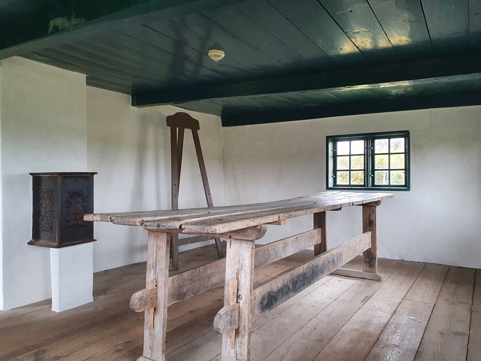 Interiør i Toftum Skole med originalt langbord, stol og jernkakkelovn under det grønmalede bjælkeloft